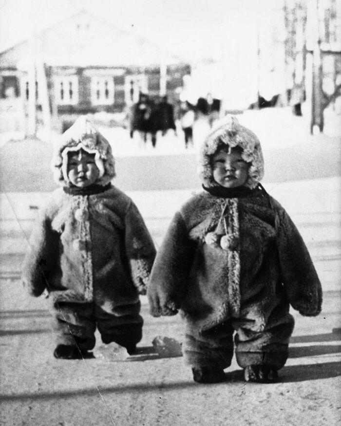 Two babies in bulky hooded outfits standing outdoors in a historical photo.