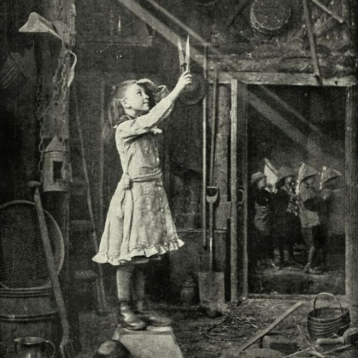 A black and white historical photo shows a young girl holding a candle, looking up, surrounded by tools in a dimly lit room.
