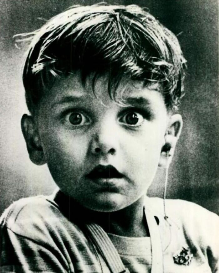 A black and white historical photo captures a young boy with wide eyes, looking surprised, with an earbud in one ear.