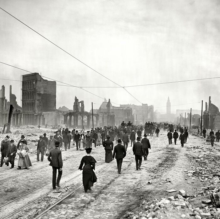 A black and white historical photo shows a wide street with many people walking amidst destroyed buildings from a disaster.