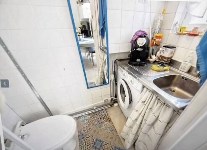 A cramped bathroom featuring a toilet, a washing machine integrated with a kitchen sink, reflecting unhinged real estate listings.