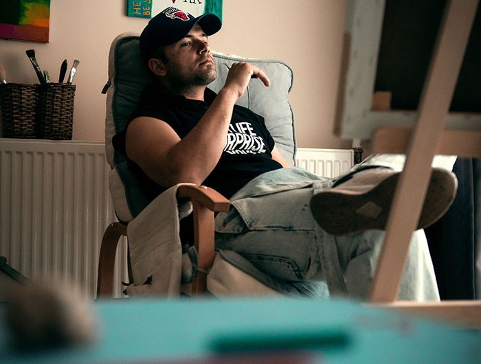 A man wearing a cap and t-shirt, relaxing in a chair, contemplating a true crime detail and realizing something is wrong.