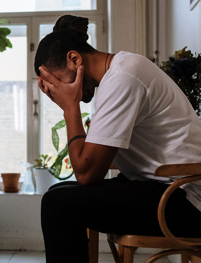 A man with dreadlocks sitting on a chair, burying his face in his hands, depicting a true crime fan's distress.