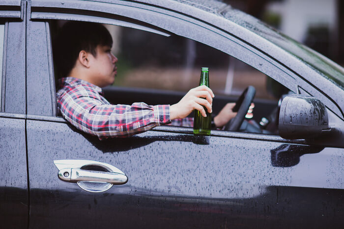 Young man in a plaid shirt holding a bottle while driving a car.