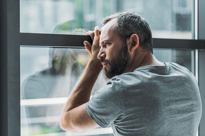 A man looking thoughtfully out a window, reflecting on secrets people keep that ruin lives.