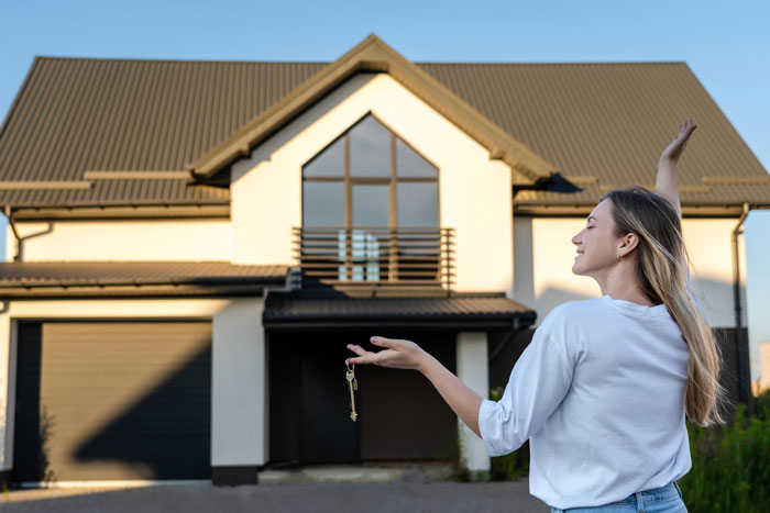 A happy woman holds keys in front of her newly bought home, celebrating her own abode.