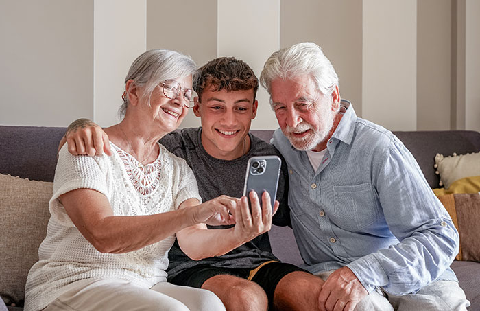 A happy 17-year-old teen with grandparents, smiling while watching something on a smartphone together.