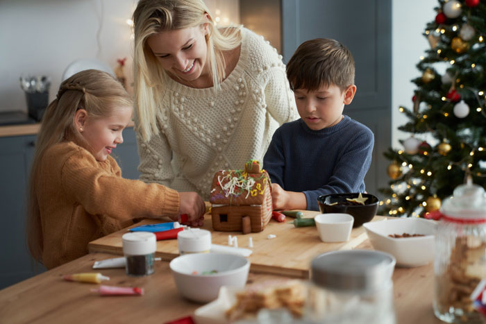 Woman and children decorating gingerbread house near Christmas tree Woman and children decorating gingerbread house near Christmas tree