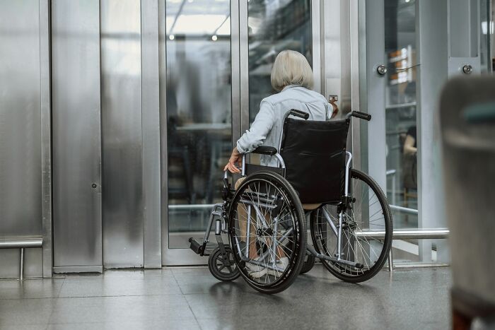 Senior woman in wheelchair waiting by elevator doors in medical facility
