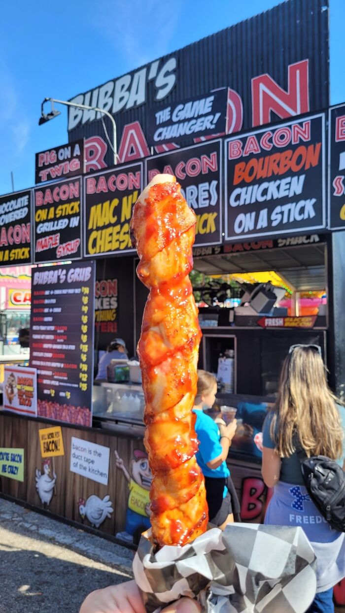 Hand holding bacon wrapped food item in front of a bacon-themed food stand