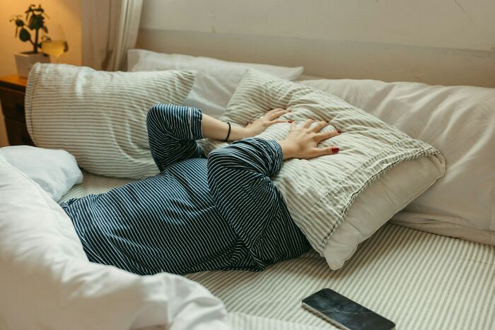 A person lying in bed with a pillow over their head, a smartphone nearby, symbolizing strange jobs or sleep.