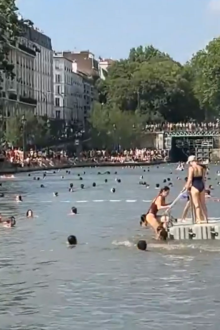 Europeans swim in a crowded river, seeking relief from the heatwave, with buildings lining the bank.
