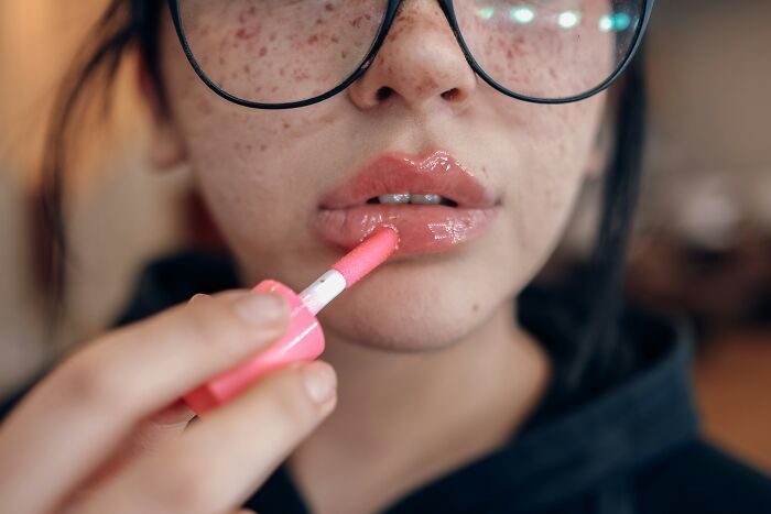 A woman with freckles and glasses applying lip gloss, illustrating a common beauty routine that can be a turn off.