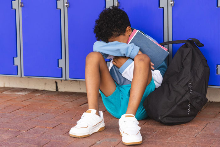 Sad child sitting by school lockers with books and backpack
