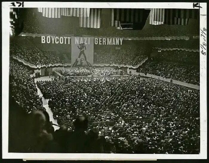 Historical photos: A vast crowd of people in a large arena under a banner that reads, BOYCOTT N**I GERMANY.