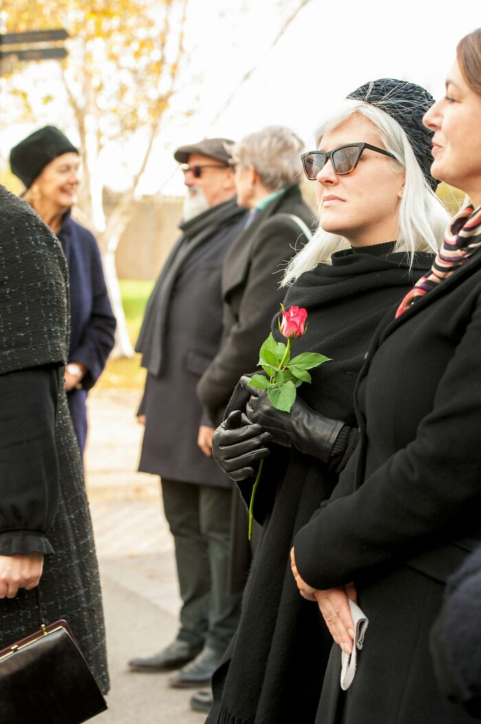 A woman holding a single red rose, dressed in black, attending an outdoor gathering where social cues are observed.