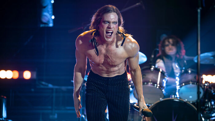 Shirtless AMC show star on stage, yelling into a microphone, with a drummer in the background, thrilling fans.
