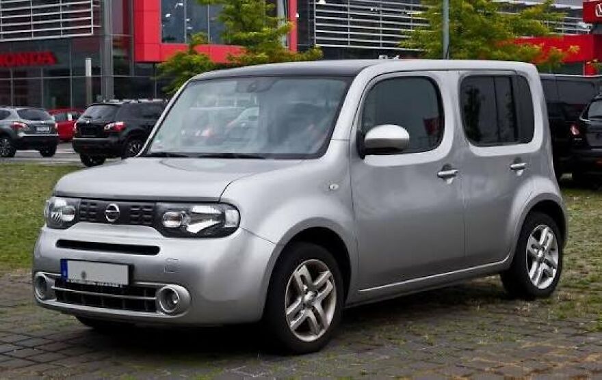 A silver Nissan Cube, an ugly car that became legendary, parked on a paved area.