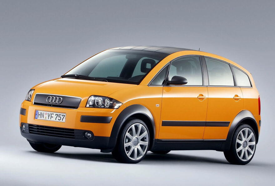 An orange Audi A2, a legendary ugly car, with a black roof against a grey background.