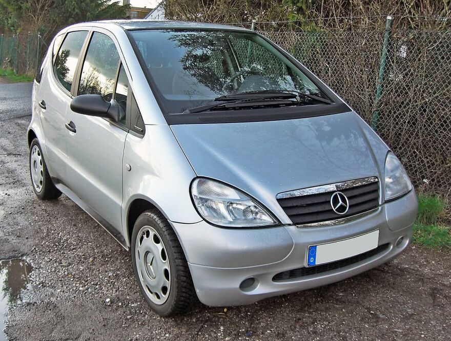A silver Mercedes-Benz A-Class, an ugly legendary car, parked on a gravel path with puddles.