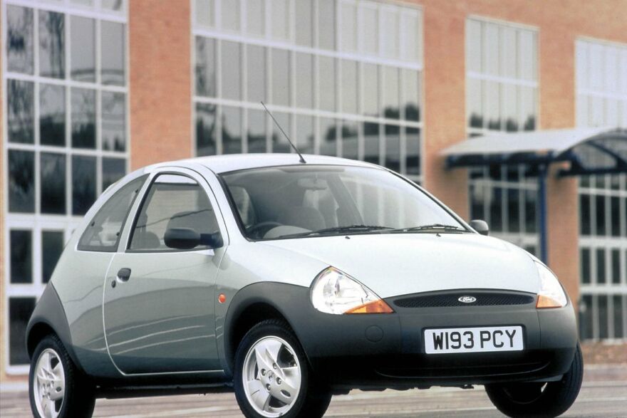 A silver Ford Ka, an ugly legendary car, parked in front of a building with large windows.