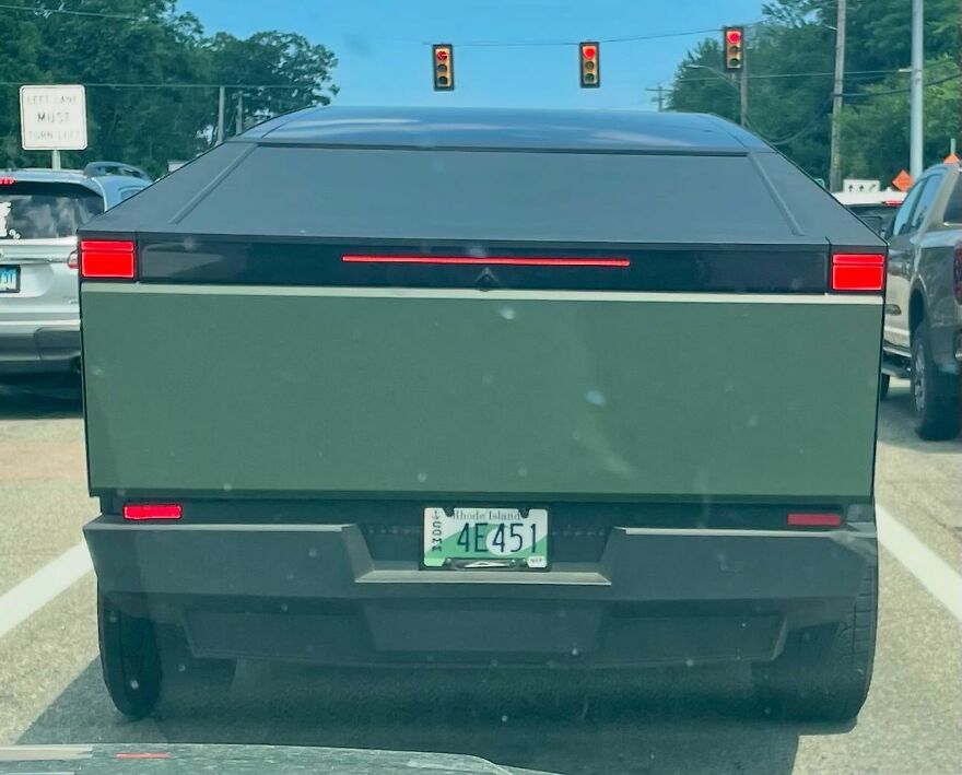 The rear view of a boxy, ugly car, possibly a Cybertruck, in green, on a road. Legendary and unique car.