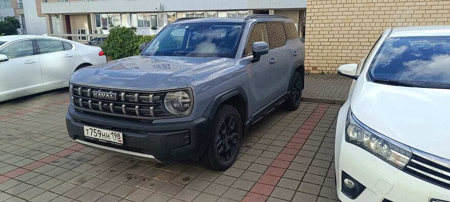 A grey Haval SUV, an ugly car that became legendary, parked in a paved lot next to a white car.