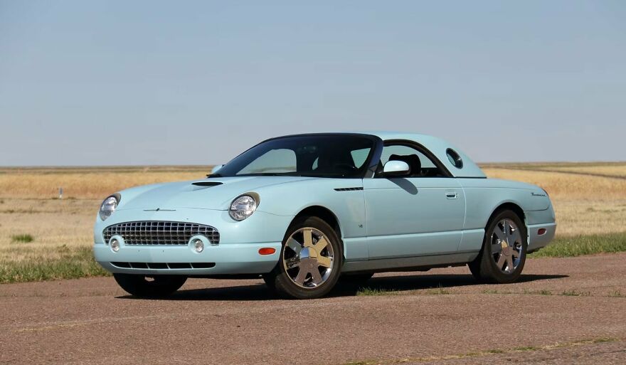 A light blue Ford Thunderbird convertible, an ugly car, parked on a paved road with fields in the background.