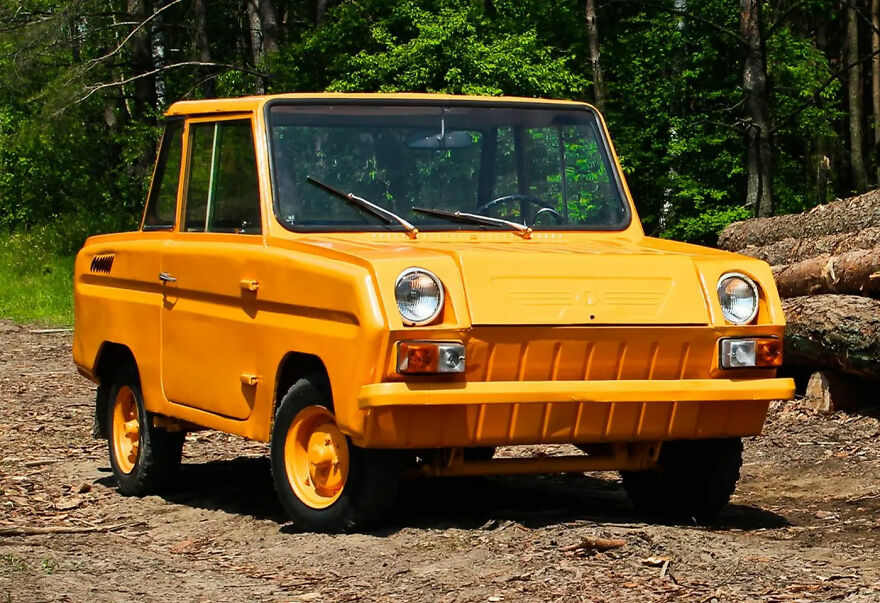 A small, boxy orange car, one of the ugly cars, parked in a forest setting with logs nearby.
