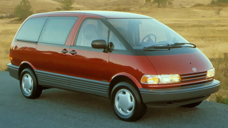 A red Toyota Previa, considered an ugly car by some, parked on an asphalt road in a rural setting. Legendary van.
