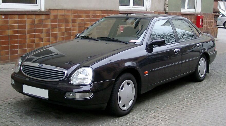A dark grey Ford Scorpio, one of the ugly cars, parked on a cobblestone street in front of a building.