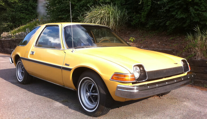Yellow AMC Pacer, a vintage legendary ugly car, parked on a paved surface with greenery in the background