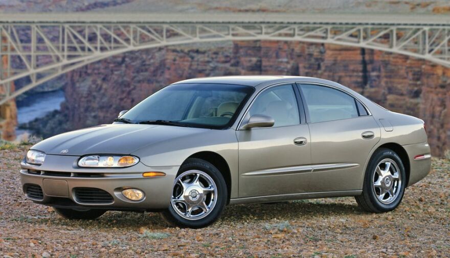 A beige ugly legendary car, a sedan, parked on a gravel path with a bridge in the background.