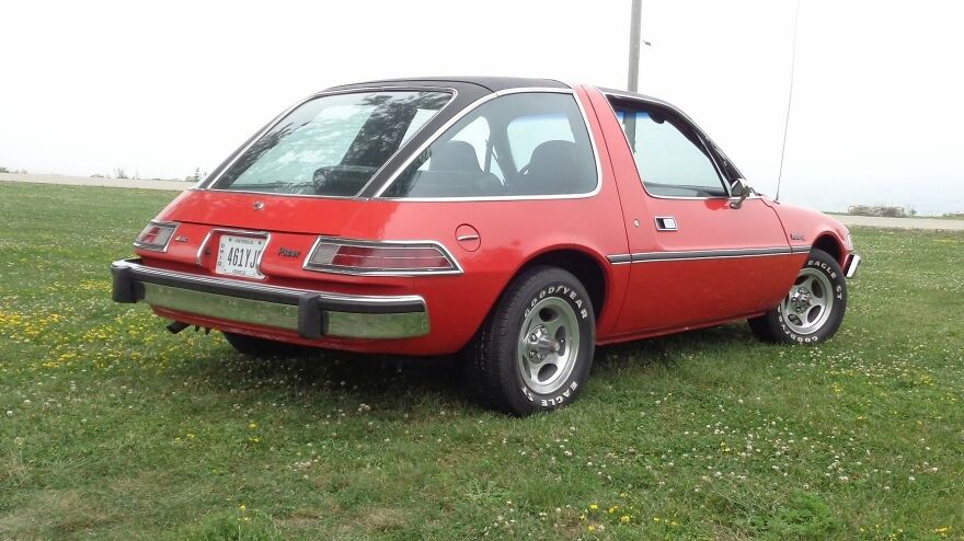 A red AMC Pacer, an ugly car that somehow became legendary, parked on a grassy field with a cloudy sky.