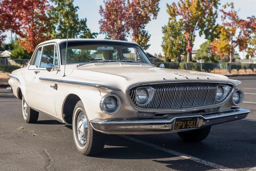 A beige Dodge Dart, an ugly car that somehow became legendary, parked outdoors with autumn trees in the background.