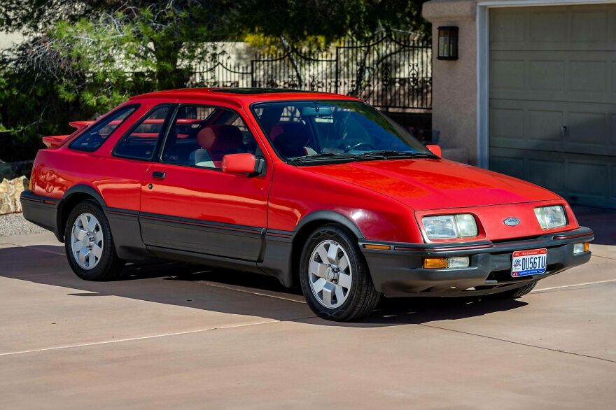A red Ford Sierra, an ugly car that somehow became legendary, parked on a paved driveway in front of a house.