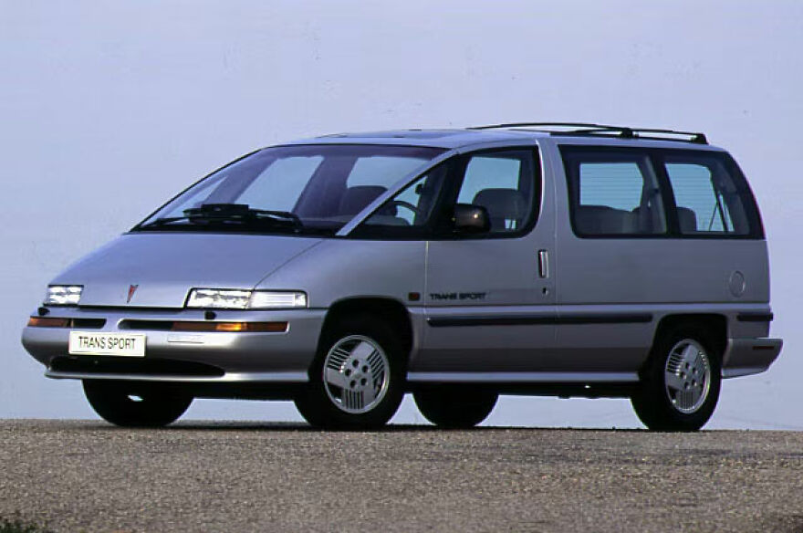 An ugly, legendary silver minivan, the Pontiac Trans Sport, parked on an asphalt road.