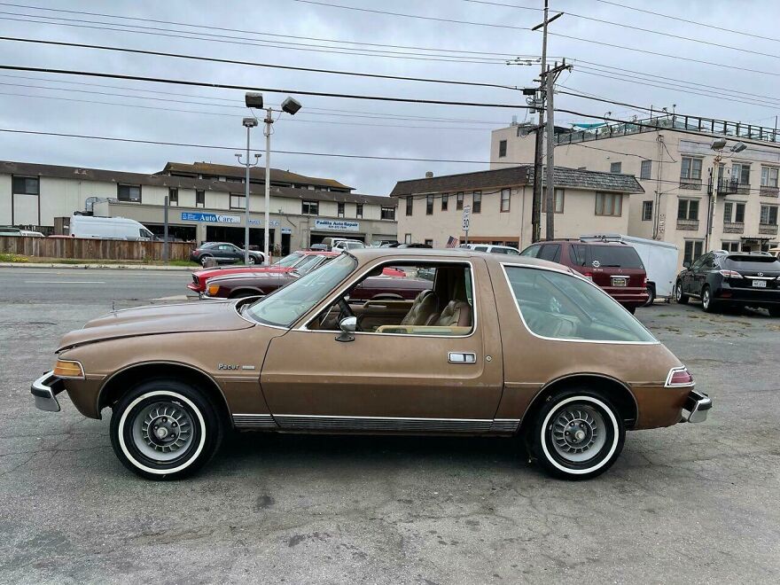A brown AMC Pacer, a legendary ugly car, parked on a street with buildings in the background.