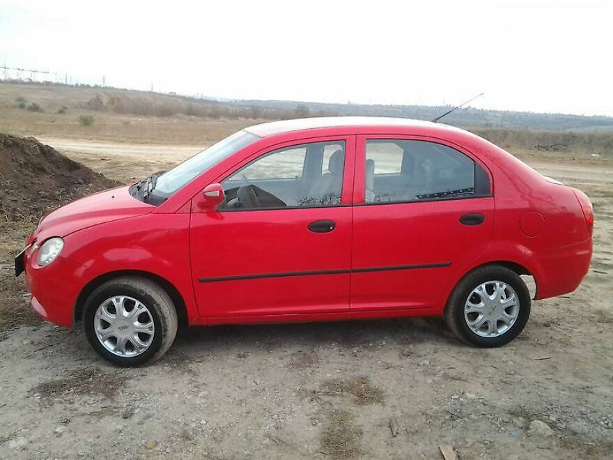 A red car, one of many ugly cars that became legendary, parked on a dirt road with a field in the background.