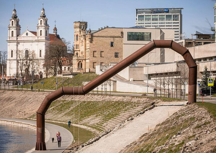 Rusty metal pipe-like monument arching over a riverside walkway