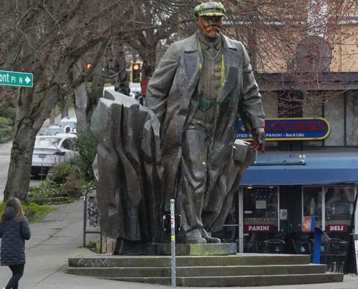 Worn metal statue of a man standing next to abstract rock in urban setting