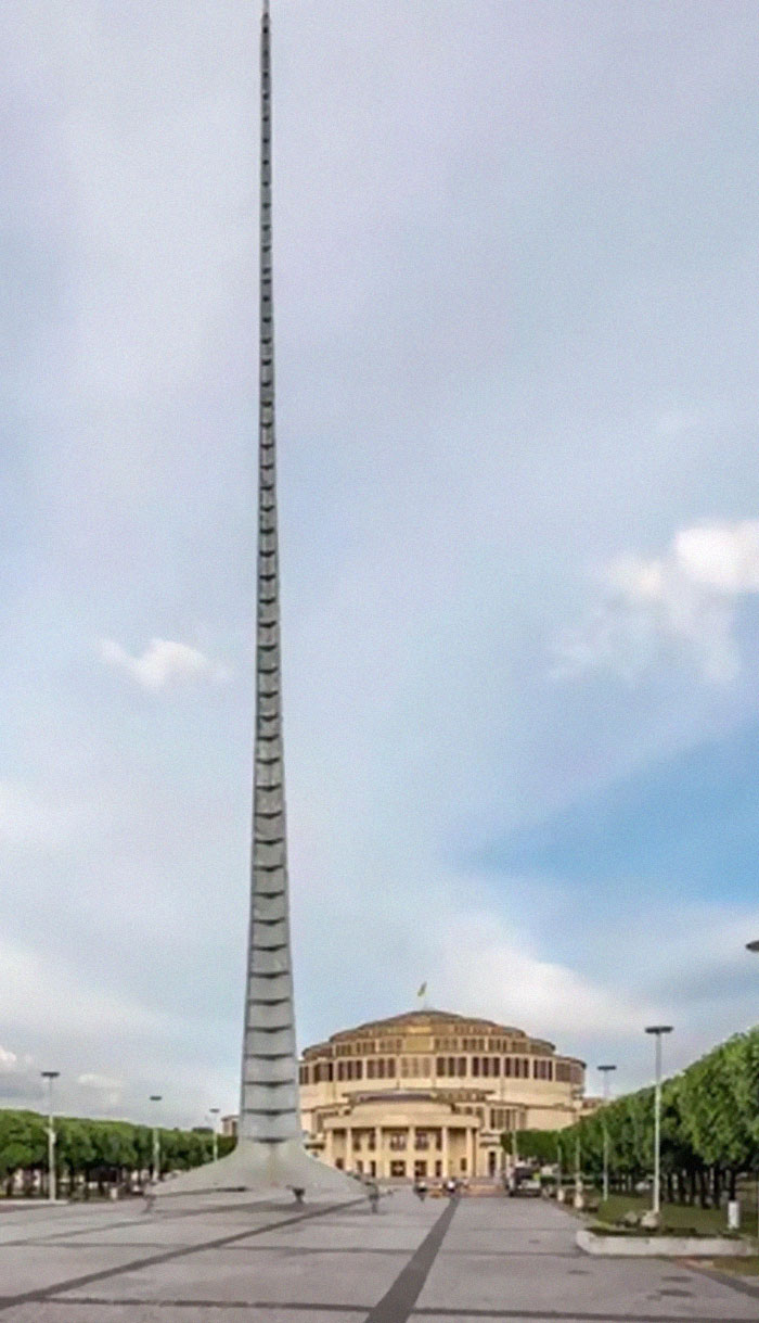 Tall, thin spire monument with round building and trees in the background