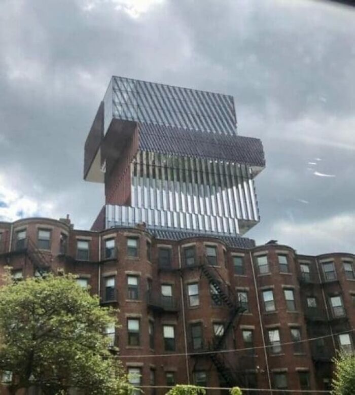 A modern, multi-story building extension appearing to balance on top of an older brick building, highlighting architecture and design fails.