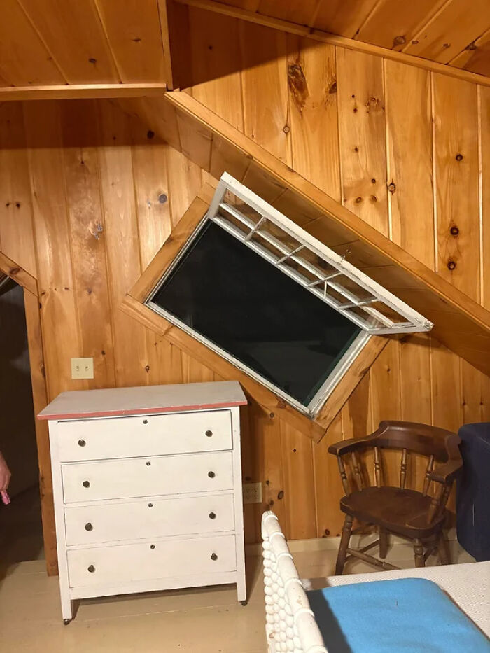 An architecture fail with a window installed diagonally in a wood-paneled room above a dresser and chair.