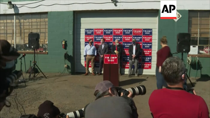 Rudy Giuliani speaks at a podium in front of a Trump 2020 banner, amidst celebrity scandals.