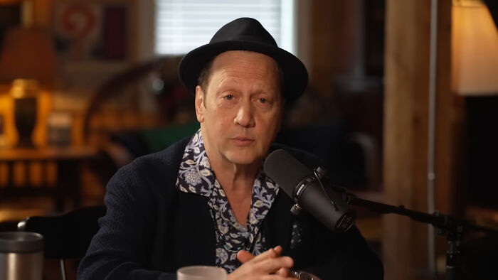 Male celebrity with a fedora, blue patterned shirt, and black jacket, sitting with a microphone, losing fame.