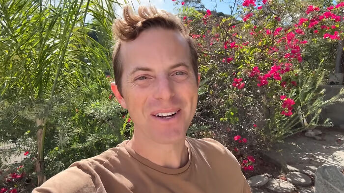 A male celebrity whose fame ran out, standing outdoors with lush greenery and pink flowers.