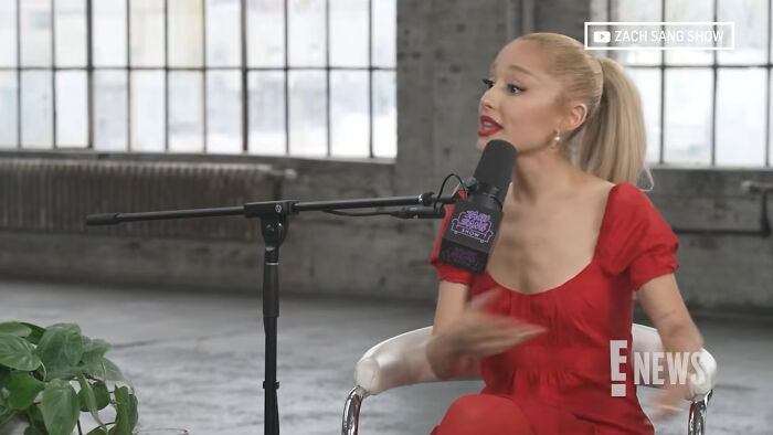 Ariana Grande in a red dress speaking into a microphone, discussing celebrity scandals.