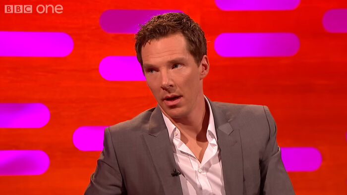Benedict Cumberbatch in a grey suit, on a show with a red and purple background, often discusses celebrity scandals.