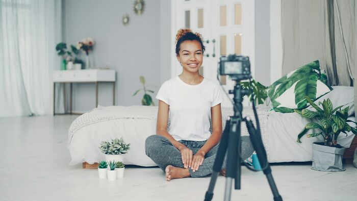 A woman sits on the floor with plants and a camera, possibly discussing health trends, intense stress and burnout.